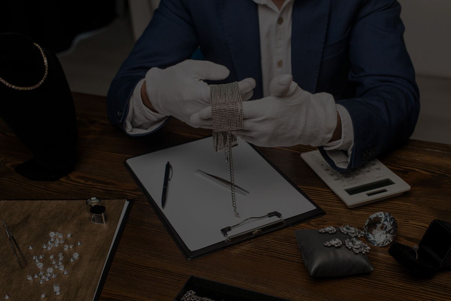 Person in a suit and gloves handling jewelry on a wooden table with tools and documents.